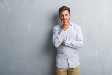 Handsome young business man over grey grunge wall wearing elegant shirt looking confident at the camera with smile with crossed arms and hand raised on chin. Thinking positive.