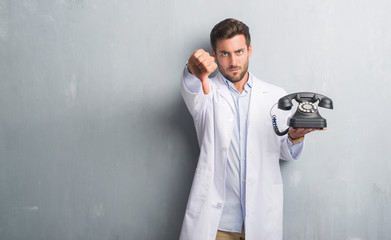 Handsome young professional man over grey grunge wall holding vintage telephone with angry face,...