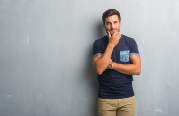 Handsome young man over grey grunge wall looking confident at the camera with smile with crossed arms and hand raised on chin. Thinking positive.