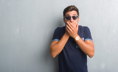 Handsome young man over grey grunge wall wearing sunglasses shocked covering mouth with hands for mistake. Secret concept.