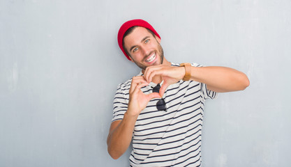 Handsome young hipster man over grey grunge wall wearing navy t-shirt and wool cap smiling in love showing heart symbol and shape with hands. Romantic concept.