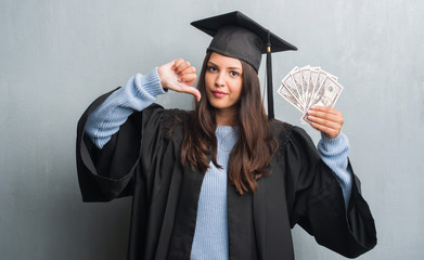 Young brunette woman over grunge grey wall wearing graduate uniform holding dollars with angry...