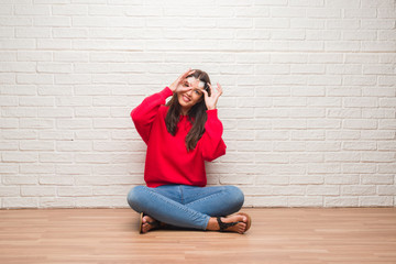 Fototapeta na wymiar Young brunette woman sitting on the floor over white brick wall with happy face smiling doing ok sign with hand on eye looking through fingers