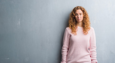 Young redhead woman over grey grunge wall wearing pink sweater with serious expression on face....