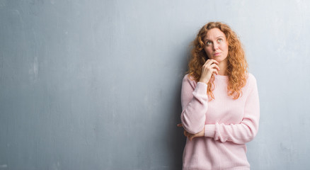Fototapeta premium Young redhead woman over grey grunge wall wearing pink sweater with hand on chin thinking about question, pensive expression. Smiling with thoughtful face. Doubt concept.