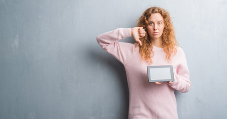 Young redhead woman over grey grunge wall using tablet with angry face, negative sign showing dislike with thumbs down, rejection concept