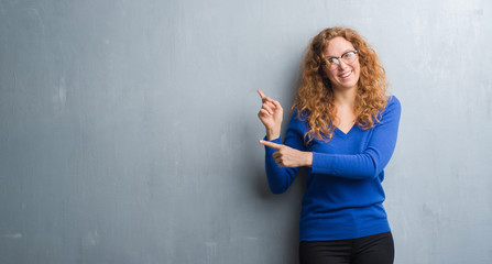 Young redhead woman over grey grunge wall smiling and looking at the camera pointing with two hands and fingers to the side.