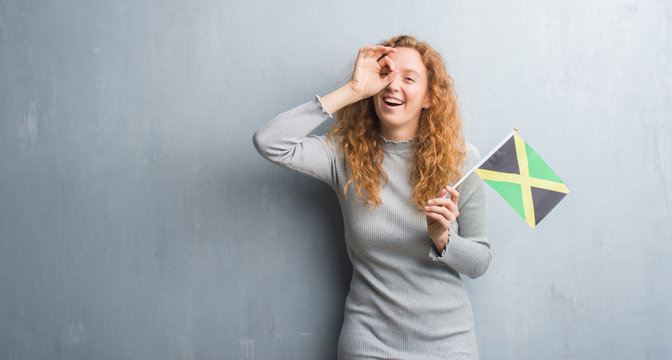Young redhead woman over grey grunge wall holding flag of Jamaica with happy face smiling doing ok sign with hand on eye looking through fingers