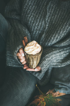 Woman in dark woolen sweater and grey jeans sitting and holding mug with hot chocolate or coffee with whipped cream and cinnamon sticks in hands. Fall warming sweet drink