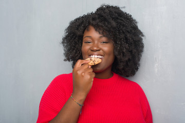 Young african american woman over grey grunge wall eating chocolate chip cooky with a confident expression on smart face thinking serious