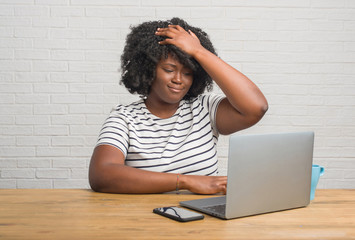 Young african american woman sitting on the table using computer laptop stressed with hand on head,...