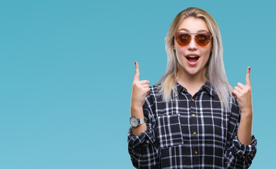 Young blonde woman wearing sunglasses over isolated background amazed and surprised looking up and pointing with fingers and raised arms.