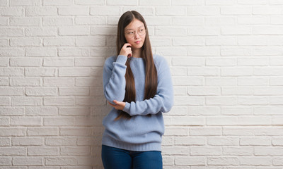 Young Chinise woman over white brick wall with hand on chin thinking about question, pensive expression. Smiling with thoughtful face. Doubt concept.