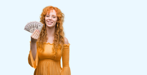 Young redhead woman holding dollars with a happy face standing and smiling with a confident smile showing teeth
