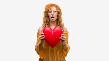 Young redhead woman in love holding red heart scared in shock with a surprise face, afraid and excited with fear expression