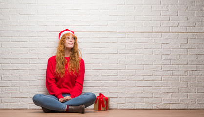 Young redhead woman sitting over brick wall wearing christmas hat smiling looking side and staring away thinking.