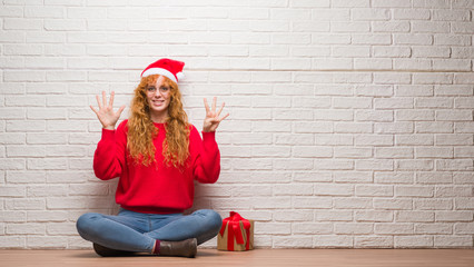 Young redhead woman sitting over brick wall wearing christmas hat showing and pointing up with fingers number nine while smiling confident and happy.