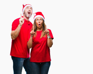 Middle age hispanic couple wearing christmas hat over isolated background amazed and surprised looking up and pointing with fingers and raised arms.