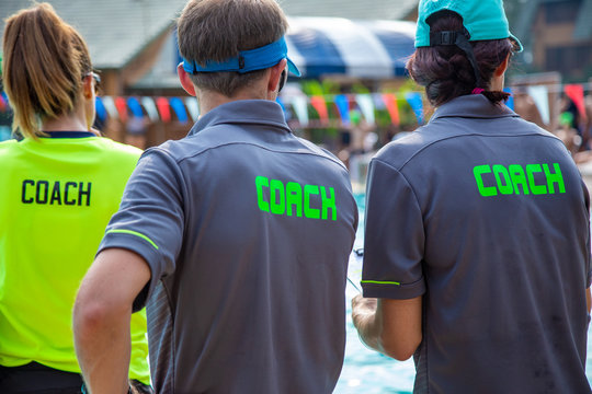 Back View Of Male  And Female Swimming Coaches, Wearing COACH Shirt, At An Outdoor Swimming Pool