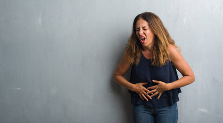 Middle age hispanic woman standing over grey grunge wall with hand on stomach because indigestion, painful illness feeling unwell. Ache concept.