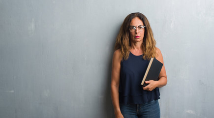 Middle age hispanic woman standing over grey grunge wall holding a book with a confident expression on smart face thinking serious