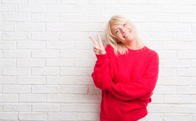 Adult caucasian woman over white brick wall wearing winter sweater smiling with happy face winking at the camera doing victory sign. Number two.