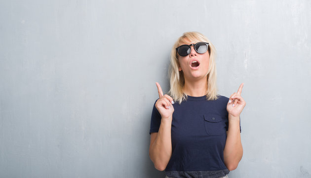 Adult caucasian woman over grunge grey wall wearing sunglasses amazed and surprised looking up and pointing with fingers and raised arms.