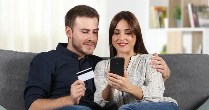 Happy couple shopping online with credit card and a smart phone sitting on a couch at home