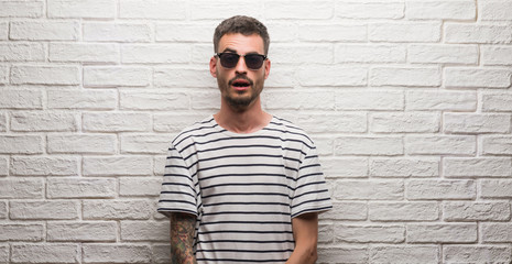 Obraz premium Young adult man wearing sunglasses standing over white brick wall afraid and shocked with surprise expression, fear and excited face.
