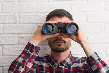 Young adult man over brick wall looking through binoculars with a confident expression on smart face thinking serious