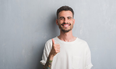 Young adult man standing over grey grunge wall happy with big smile doing ok sign, thumb up with fingers, excellent sign