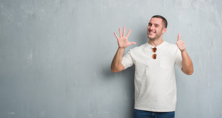 Young caucasian man over grey grunge wall wearing sunglasses showing and pointing up with fingers number six while smiling confident and happy.