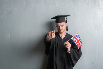 Young redhead man over grey grunge wall wearing graduate uniform holding united kigdom flag pointing with finger to the camera and to you, hand sign, positive and confident gesture from the front