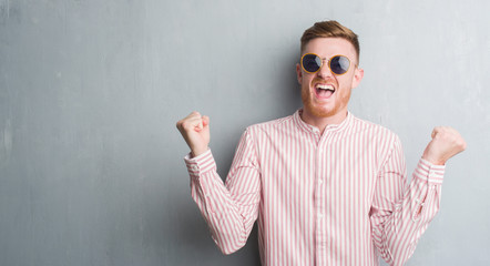 Young redhead man over grey grunge wall wearing retro sunglasses screaming proud and celebrating victory and success very excited, cheering emotion