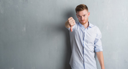 Young redhead business man over grey grunge wall looking unhappy and angry showing rejection and negative with thumbs down gesture. Bad expression.