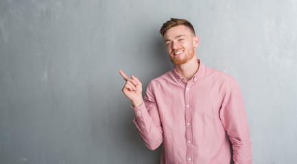 Young redhead man over grey grunge wall wearing pink shirt with a big smile on face, pointing with hand and finger to the side looking at the camera.