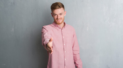 Young redhead man over grey grunge wall wearing pink shirt smiling friendly offering handshake as greeting and welcoming. Successful business.