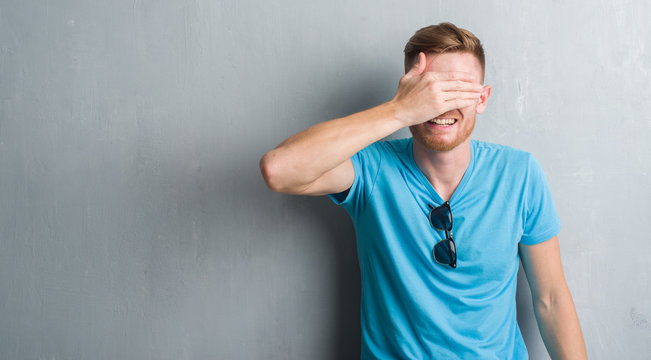 Young redhead man over grey grunge wall wearing casual outfit smiling and laughing with hand on face covering eyes for surprise. Blind concept.