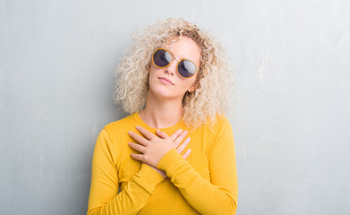 Young blonde woman with curly hair over grunge grey background smiling with hands on chest with closed eyes and grateful gesture on face. Health concept.