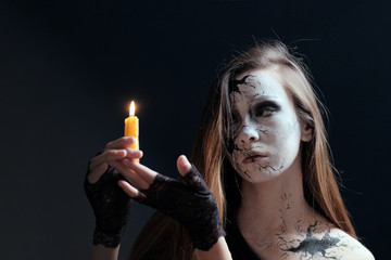 Obraz premium Makeup in the style of Halloween. A young girl with long hair with cracks painted on her face is holding a burning candle. Dark background. Copy space. Black dress. The image of the witch or doll.