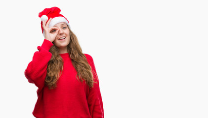 Young blonde woman wearing santa claus hat with happy face smiling doing ok sign with hand on eye looking through fingers
