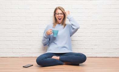 Beautiful young woman sitting on the floor driking cup of coffee at home annoyed and frustrated shouting with anger, crazy and yelling with raised hand, anger concept