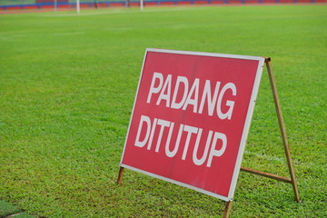 Sign on fields titled "PADANG DITUTUP" on green grass inside a stadium