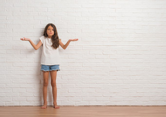 Young hispanic kid stading over white brick wall clueless and confused expression with arms and hands raised. Doubt concept.