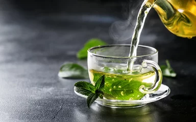 Fotobehang Thee Hete chinese groene thee met munt, met plons die uit de ketel in de beker stroomt, stoom stijgt, donkere achtergrond, selectieve focus