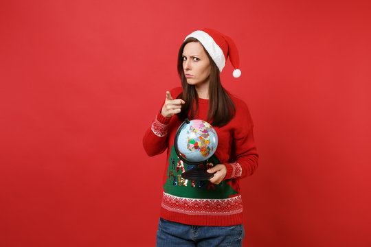 Strict young Santa girl in Christmas hat holding world globe pointing index finger on camera isolated on red wall background. Happy New Year 2019 celebration holiday party concept. Mock up copy space.
