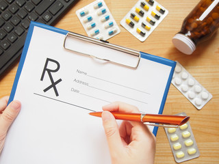 Female doctor hands holding clipboard and writing prescription or rx form to patient on office desk with keyboard and medicine tablet. Top view of doctor's desk. Healthcare and medical concept.