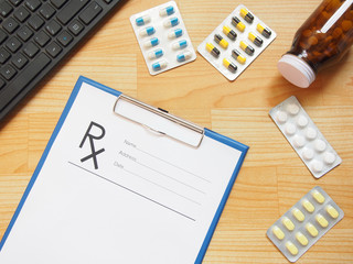 Blank prescription on a blue clipboard with pen, keyboard and medicine all around on wooden table. Top view of doctor's desk with copy space. Healthcare and medical concept.