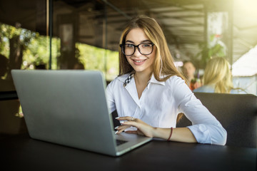 Taking advantages of free Wi-Fi. Beautiful young woman in glasses working on laptop and smiling while sitting outdoors in cafe