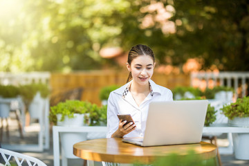 An attractive young businesswoman using her phone and laptop at a cafe outdoors